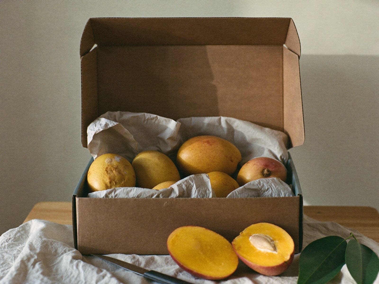 An open kraft box of ripe mangoes beside a halved fruit in soft morning light