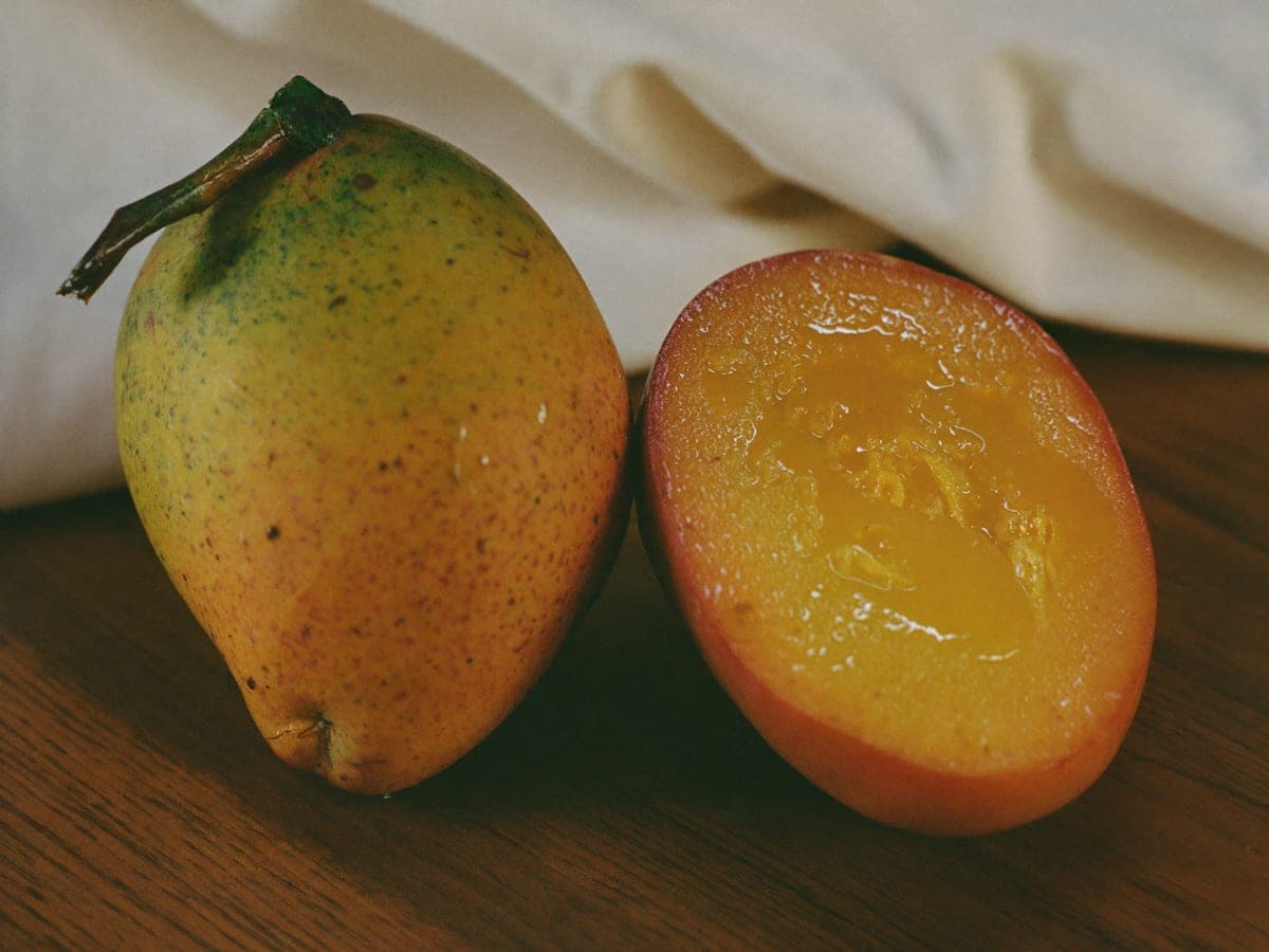 Two Chaunsa mangoes on dark wood: one whole with a green shoulder blushing through yellow to red, the other halved to reveal deep orange, glossy, fibre-free flesh.
