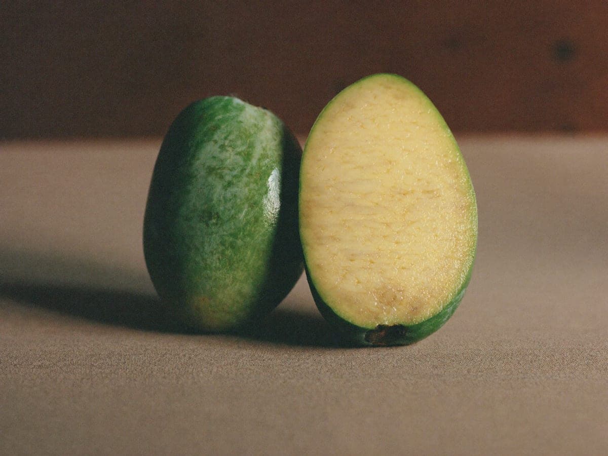 Two Langra mangoes on tan cloth: one whole and still deep green although fully ripe, the other halved to show pale yellow-green flesh running right to the skin.
